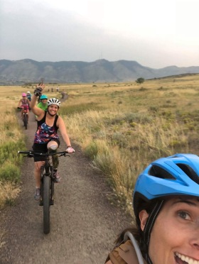 Lauran taking a selfie with me waving while riding in the background. Other women on mountain bikes are behind me.